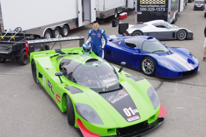 The Best looking Kit Cars - Page 11 - Kit Cars - PistonHeads - The image captures an exhilarating racing scene on a dirt road, where two high-performance racing cars, painted in vibrant hues of green and blue, are prominently featured. The green car is on the left side of the photo, while the blue car is parked on the right. Notably, a person is standing next to the blue car, adding a human element to the scene. The background is filled with a line of trailers, hinting at the ongoing activity or a potential race route. It appears to be a professional or organized racing event, given the location and the preparation of the cars.