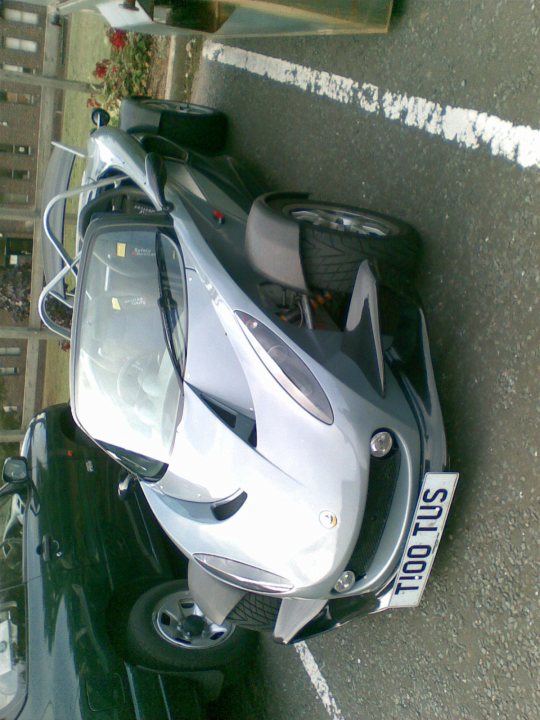 Anyone know what this Lotus is? - Page 1 - General Lotus Stuff - PistonHeads - The image captures a light gray or silver sports car positioned on the passenger side, facing left. It is parked along a white line on what appears to be an asphalt surface. In the background, there are green plants, and a portion of another vehicle can be seen to the left of the sports car. The vehicles seem to be parked in an urban setting.