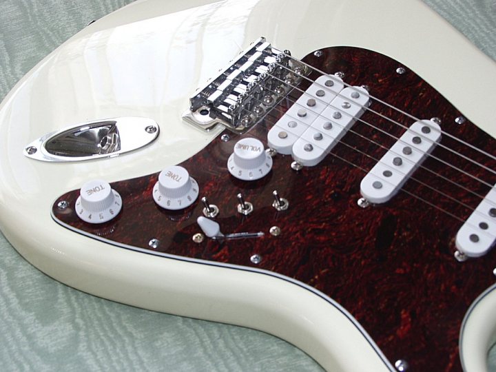 Pistonheads Luthiers - The image showcases a white electric guitar with a wooden neck, exhibiting a fusion of white with a rosewood finish. The guitar sports two pickups, both hooked up to a pickguard that has a distinct shape. One of the pickups has a silver cover, while the other has a creamy white one. The white guitar strings clad the pickguard, completing this modern electric guitar setup.