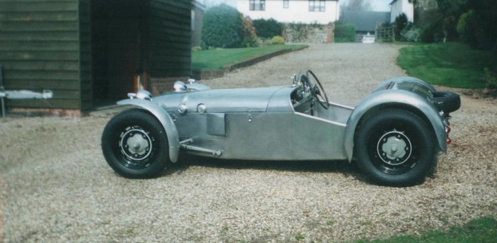 Tribute To A Racer - Page 1 - Classic Cars and Yesterday's Heroes - PistonHeads - The image features an antique sports car parked on a gravel driveway. The car has a classic design with a vintage look, including a curved body, prominent headlights, and wire wheels. It appears to be deep or sickly silver in color, contrasting with a black/brown roof section. There is a person sitting in the driver's seat, but they are not fully visible, and the photograph seems to have been taken at a slight angle. The setting is outdoors with a residential background, showcasing a quaint house and lush greenery, under what might be a partly cloudy sky, suggesting it could be early morning or late afternoon. There are no distinguishable texts or logo visible in the image to provide additional context.