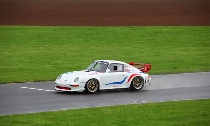 Pistonheads - The image captures an exhilarating moment on a car track. A white Porsche race car, adorned with blue and red stripes, is the star of the scene. It's speeding around an immaculate green circuit, leaving a trail of water spraying behind on the tarmac. The Porsche is in motion, heading towards the right side of the image, its driver focusing intently on the race. The background of the image is a blur, emphasizing the car's swift motion and the adrenaline rush of the race.