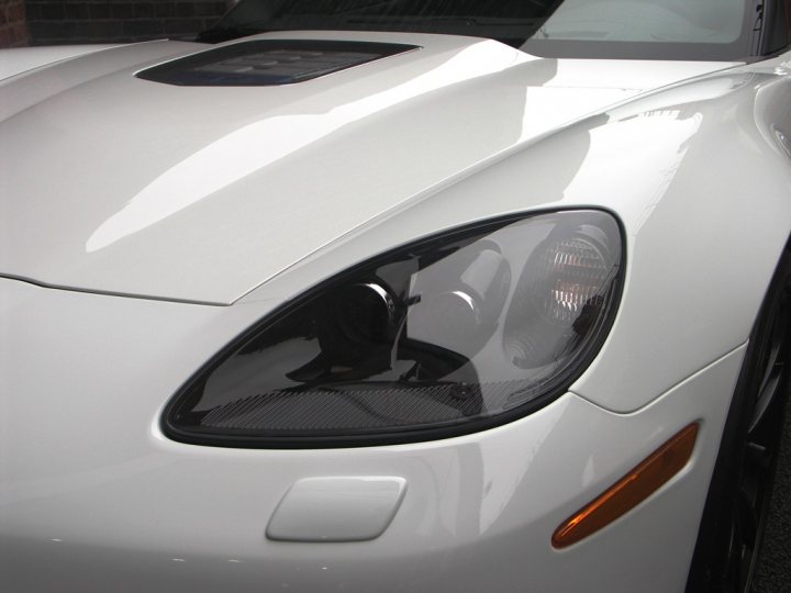 How about some ZR-1 photos to cheer you up? - Page 1 - Corvettes - PistonHeads - The image shows a close-up view of the front of a white car with a shiny finish. The car appears to be a modern sports or luxury vehicle, as suggested by the aggressive aerodynamic design and the sleek headlights. The headlight assembly includes separated turn and off-road lighting components. There is a prominent grille area at the front, which could indicate an air intake or be used for decorative purposes. The car is currently parked, but there is a slight reflection of the photographer on the vehicle. The setting seems to be in a garage or covered parking area due to the absence of visible surroundings.