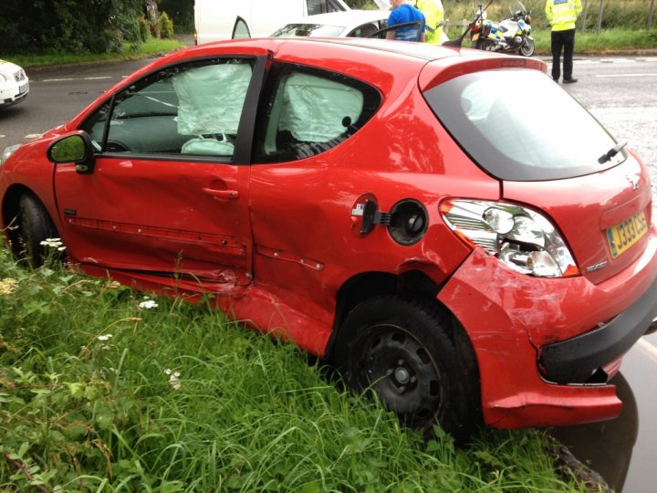 End of Peugeot.. - Page 2 - General Gassing - PistonHeads - The image presents a scene of a red car damaged on its side, sitting on the side of a road. The car is quite prominent, and its damaged side indicates it has been in some sort of accident. There are no visible people near the car, but several motorcycles can be seen in the background, hinting at possible onlookers or the site of an accident. Surrounding the car is sparse vegetation, including grass and some small plants, indicating that the car has likely been there for a while, as nature is slowly reclaiming the area. The presence of a person wearing a blue jacket and khaki pants on the right side of the image adds a human element to the scene.