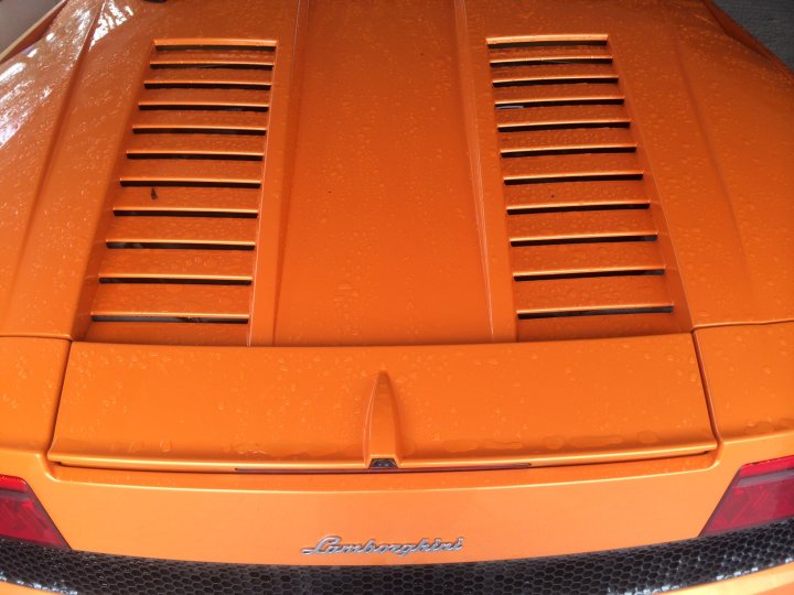 More pics of my LP560 - Page 1 - Gallardo/Huracan - PistonHeads - The image shows a portion of an orange car, specifically focusing on the tail end. The car features a black diffuser that spans its width, accentuating the rear view. The trunk of the car is visible, with the name "Lamborghini" etched onto it, reflecting the brand of the vehicle. Several vents are positioned across the trunk and the area above it, possibly for aerodynamic purposes. The area is framed by a black element, adding a contrast to the car's orange body.