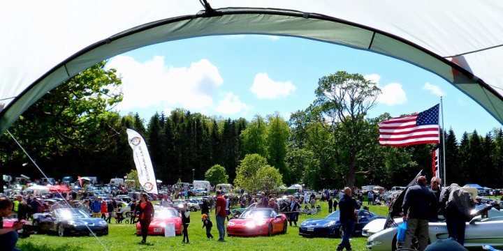 Thirlestane Castle Classic Car Show - Page 1 - Scotland - PistonHeads - This image captures a lively scene at a car show. The event is held in a park-like setting, where rows of vintage cars, including a few sports cars and a truck, are parked. The cars include a notable classic red convertible. Among the crowd, there's a man holding an American flag, perhaps indicating a patriotic event or a celebration of a distinct type of automobile, such as classic American muscle cars. The scene is bustling with people, all seemingly in high spirits, walking amongst the parked cars. An overhead tent provides shade and a backdrop to the event. The overall atmosphere seems vibrant and community-oriented, creating a sense of nostalgia for the golden age of automobile culture.