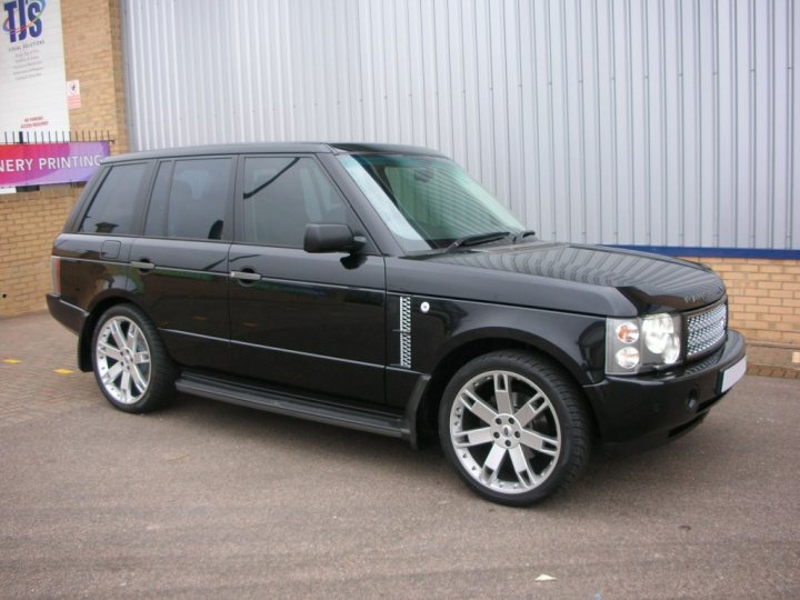 Pistonheads Rover Range Sell - The image depicts a black Range Rover parked on a concrete surface. The vehicle features the distinctive off-road styling of the Range Rover brand, including large wheels and a prominent body shape. There are three visible doors on the vehicle, and a spare tire is mounted underneath the back of the vehicle. In the background, part of a building can be seen, suggesting this location may be a parking area near a commercial or industrial building. The lighting in the photograph indicates it may not be daytime.