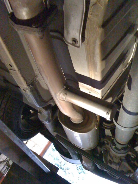 What's this? - Page 1 - Honda - PistonHeads - The image appears to be a close-up photograph of a vehicle's underside. Visible components include an exhaust pipe with a split front, a metal protective cover, and a muffler or exhaust to the right. The focus of the image is on the metalwork, showcasing the vehicle's mechanical components. The upper boundary of the image is dominated by a concrete floor, and there's natural light coming from the upper left corner, suggesting the photo may have been taken outside. The style of the image looks casual and industrial, capturing a moment of vehicle maintenance.