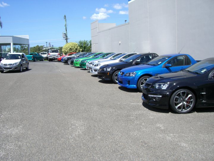 Wheres Stevo Pistonheads - The image shows a parking lot with several cars parked in a straight line along the wall. The cars vary in color, predominantly navy blue, black, and a few in white and red. They are all standing upright, presumably for photos as one car has a notable upturned hood. The parking lot appears to be in an industrial or commercial area, and a large building forms the backdrop of this scene. The setting suggests it might be a sunny day, possibly indicating the start of a business or car dealership day.