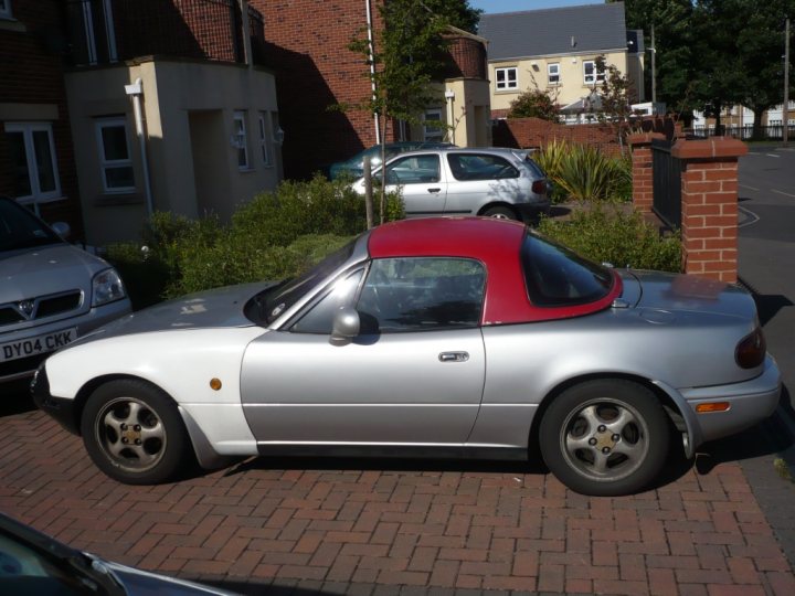 Booked Bodyshop Pistonheads - The image depicts a single-occupant convertible car parked on a brick paved driveway. The car has a unique red top and a red rear windshield. The vehicle's hood and front fenders are in a contrasting white color, creating a striking visual effect. In the background, other vehicles and buildings can be seen, suggesting that the photo was taken in an urban or suburban setting. The car appears to be in good condition, which is consistent with its well-maintained exterior.