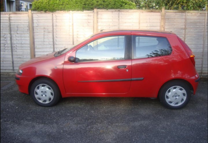 Pistonheads - The image shows a red hatchback parked outdoors on an asphalt surface. The car has a glossy finish, and its reflection is visible on the surface next to it. There's a suburban fence in the background, and lush green foliage is across from the parking area, suggesting a residential neighborhood. The photo appears to be taken during daylight with ample natural light illuminating the scene.