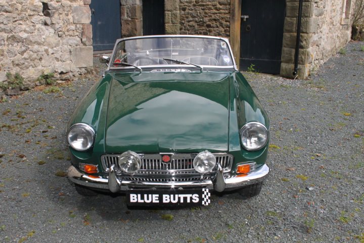 Thoughts On This £12K MGB - Page 1 - MG - PistonHeads - The image features a green vintage sports car parked on a gravel driveway. The car's convertible top is down, and it has a classic design with two headlights and round taillights. There is a sign on the back of the car stating "Blue Butts," likely indicating the vehicle's registration or ownership. The setting appears to be in a rural or semi-rural area, with a stone wall visible on the left side of the image, suggesting an old, possibly historic, building nearby. The car's condition and presence of the "Blue Butts" sign suggest it might be participating in an informal meet-up or event for classic or sports cars.