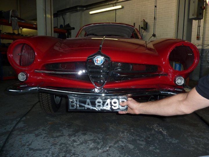 Restoration pics - Page 6 - Classic Cars and Yesterday's Heroes - PistonHeads - The image shows a person in a black shirt holding a vehicle registration plate from a vintage or classic car. The car itself has a sleek design with red fenders and a hood ornament, reminiscent of European vintage automobiles. The registration plate bears the letters "BLA", with the numbers "849" underneath it. The setting appears to be an indoor garage or workshop, as indicated by the presence of what might be a wire or conduit on the right side of the image and the overall ambiance.