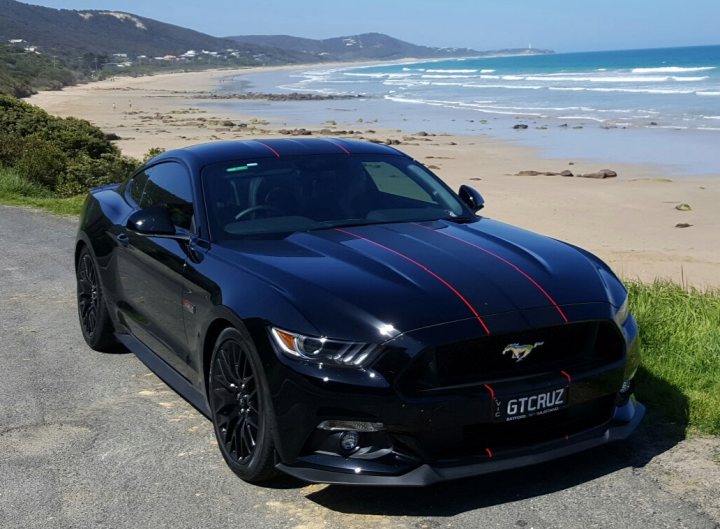 Real Good Number Plates vol 5 - Page 34 - General Gassing - PistonHeads - The image portrays a dynamic black Mustang sports car parked amidst a picturesque beachfront scene. The car, positioned on the left side of the frame, boasts a striking exterior, accented with red stripes running the length of the hood. Its glossy finish and the hood's compact lines suggest a model with a blend of speed and sophistication.

Behind the car, the tranquil beach evokes a sense of serenity. The light gray color of the sand contrasts nicely with the deep blue of the ocean, while the clear sky, visible in the background, adds to the overall beauty and serenity of the scene. The noises from the ocean are the only sounds in the otherwise peaceful setting, creating a harmonious balance between nature and man-made beauty.