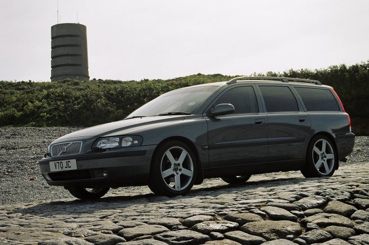 Pistonheads - This image features a gray SUV parked on a gravel surface with a backdrop of a clear sky. The vehicle appears to be modern and is positioned slightly off-center in the frame. Behind the SUV, there is an old water tower, which adds a contrasting element to the more contemporary car. The lighting suggests it is either early morning or early evening, given the soft, diffused light casting shadows on the ground.