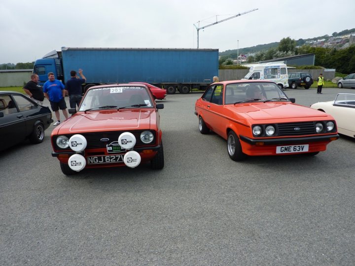 Pistonheads West Classic Wales - In this image, two classic orange cars are parked side by side on a blacktop surface within what appears to be a spacious parking lot. The car on the left is equipped with a white light bar, possibly for police or emergency use. Behind the cars, several other vehicles, including a truck, are visible, suggesting a larger gathering or event in the background. The weather seems to be overcast, with gray skies above the scene.