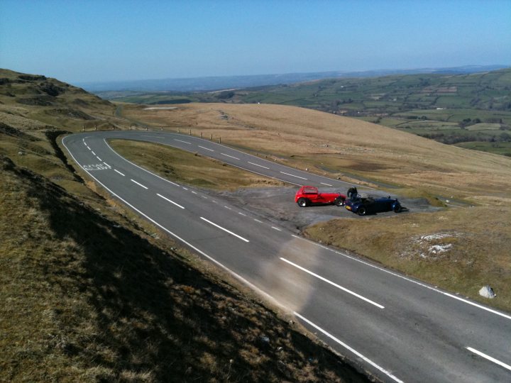 Blat Pistonheads Wales - The image captures a serene rural landscape featuring a winding highway. At the bottom of a small hill embracing both sides of the road, an old red car and a black truck are parked. The road itself is curved and marked by white and yellow stripes. Above, the sky is clear, accentuating the vastness of the area dotted with a few distant hills. The image exudes a sense of solitude and tranquility.