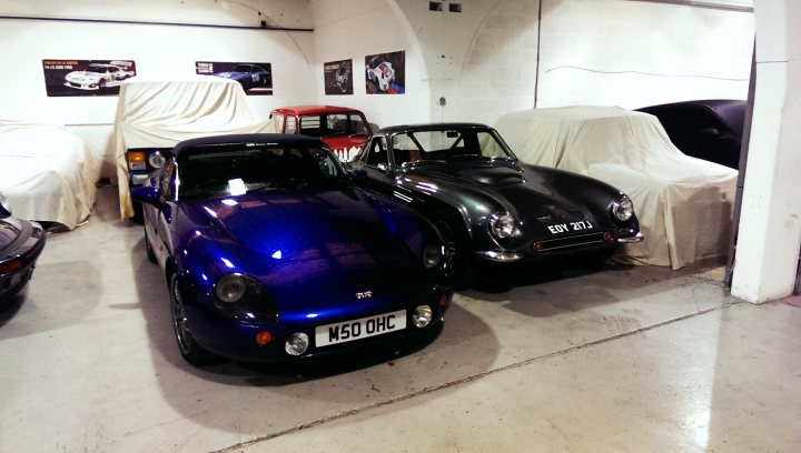 Random TVR pics - Page 409 - General TVR Stuff & Gossip - PistonHeads - The image captures a garage filled with vintage cars, their black, blue, red, and maroon bodies reflecting the ambient light. Some of these cars are covered, hinting at the care and attention given to their preservation. The space is confined, with the cars parked closely together, creating a scene reminiscent of a time gone by. The garage itself is simple, with white walls and a solid floor, allowing the colors of the cars to stand out. A few posters and a shelf with a car part on it are visible, suggesting the hobby and possibly the passion of the owner for these classic vehicles.