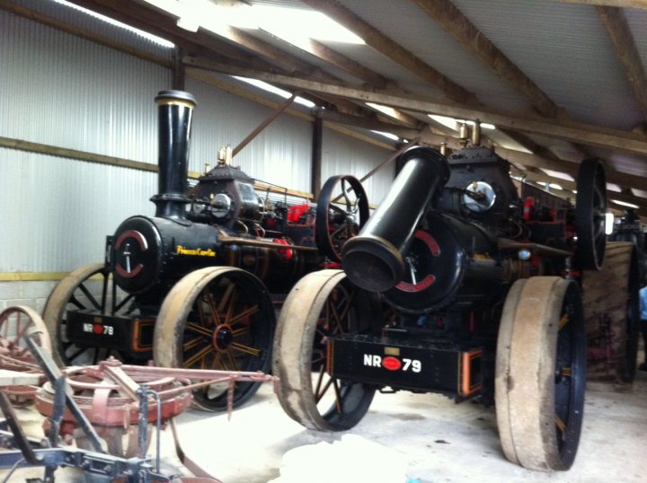 Garage Queens - Page 10 - Aston Martin - PistonHeads - The image features a vintage steam locomotive, possibly an attraction for museum visitors. The train is black and prominently displayed inside a building with a corrugated metal roof. Multiple steam pipes are visible, indicating the locomotive's sturdy build and the era it represents. It's parked in front of a chain link fence, suggesting it could be located in a yard or a repair facility. There is also a plow in the vicinity of the locomotive, which might be used for vintage vehicles restoration or maintenance. The overall setting appears to be an industrial or historical environment.