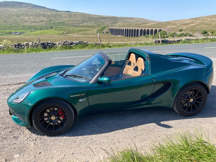 The big Elise/Exige picture thread - Page 61 - Elise/Exige/Europa/340R - PistonHeads UK - The image shows a green sports car parked on what appears to be the side of a road. It's positioned at an angle, allowing a clear view of its sleek design and black wheels. The vehicle is parked next to a grassy area and there are no other cars visible in the vicinity. In the background, there's a hint of a building structure with a white facade and some greenery. The overall scene suggests a rural or semi-rural setting.