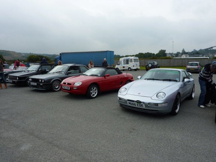 West Pistonheads Wales Classic - This image appears to captures a scene at an outdoor event or gathering involving a group of individuals and several parked vehicles. The vehicles vary in size, with models like a dark hatchback, a red sports car, and a light-colored convertible. They are stationed on a paved surface, and people are scattered around them, suggesting socializing or shared interest in the cars. The background is somewhat nondescript, featuring a clear sky and some greenery, as well as partial views of other vehicles and small buildings or structures which are not in focus.