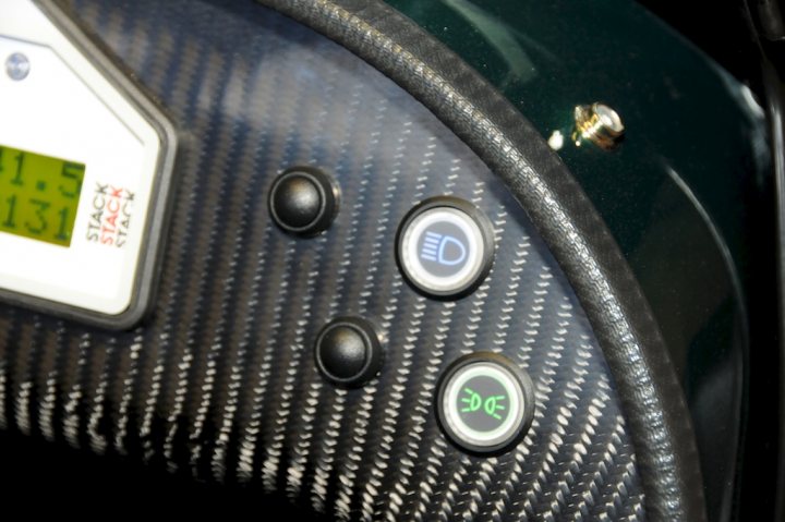 New Style Dash Switches on my R500 - Page 1 - Caterham - PistonHeads - The image showcases a close-up view of a vehicle's steering wheel with a carbon fiber texture. The wheel features three black buttons, each situated near the 12:3 position on the wheel. To the left, there's a smaller panel with a digital display indicating the time '11:13'. The entire setup suggests that this is the driver's side of a luxury or sports car. The steering wheel's design and the presence of the digital display hint at advanced technology and a high-end interior.