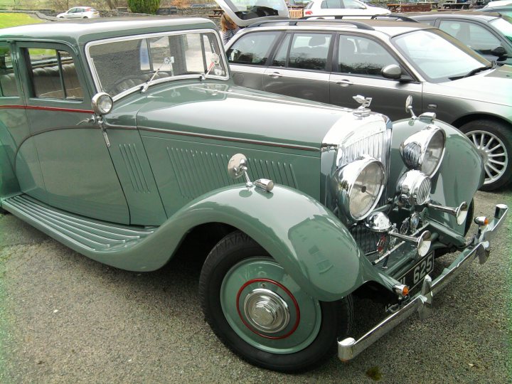 Saw this today... - Page 1 - Classic Cars and Yesterday's Heroes - PistonHeads - This image captures a scene in a parking lot where two vintage cars, specifically pre-WWII era ones, have been parked. The showcase car is an older green sedan with distinctive features such as a narrow front grille, heart-shaped side mirrors, and a spare tire on the back. It is facing the camera but seems to have an object obstructing the view of its front license plate. Around it, there are other cars parked, slightly obscured by the front end of the main subject. The setting has a car wash visible in the background along with a patch of trees and what appears to be a building.