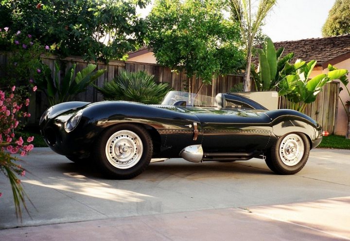 Other cars you envy? - Page 1 - Aston Martin - PistonHeads - In the foreground of the image, an impressive black racer car stands on a driveway, gleaming under the sunlight. The car's sleek design and shiny black exterior highlight its aerodynamic shape and imposing size. The surrounding area is accentuated with tropical plants and vibrant flowers, providing a stark contrast to the masculine presence of the car. The scene exudes a sense of luxury and leisure.