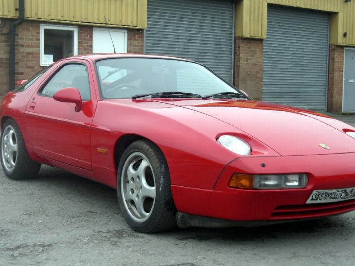 A red and black truck parked in a parking lot - Pistonheads - The image shows a vibrant red Porsche sports car parked in what appears to be an outdoor parking area, situated in front of a simple, metal gray garage door. The car features a sleek and aerodynamic design, characteristic of sports cars, with the distinctive Porsche logo on the front. The license plate on the car reads 'DJ59 A', providing a unique identifier. The vehicle has alloy wheels, and the overall setting suggests a casual, everyday scene rather than a staged or promotional event.