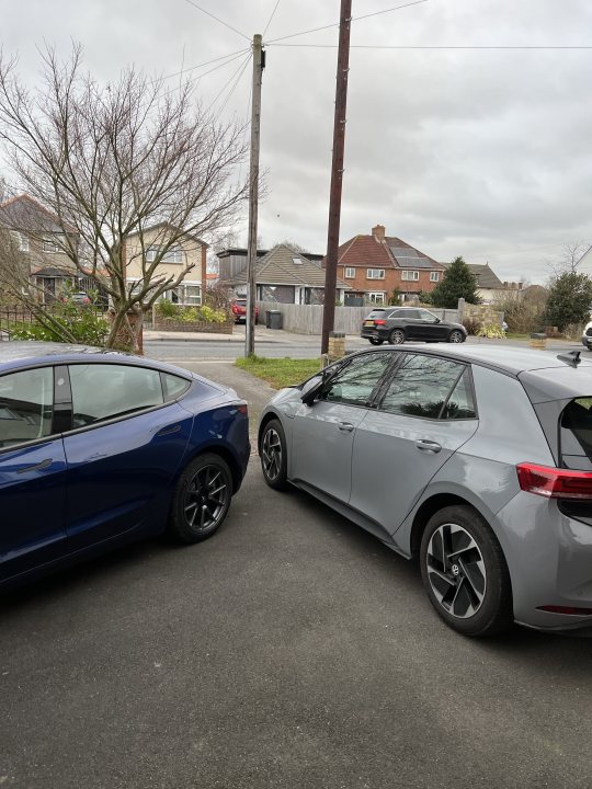 E-Golf to Tesla Model 3 - am I mad? - Page 2 - EV and Alternative Fuels - PistonHeads UK - The image is a photograph taken outdoors during the day. It features two cars parked side by side on what appears to be an asphalt area, possibly a parking lot or street. The car in the foreground is blue and has its doors open, revealing interior details. Next to it is a silver car, also with its door open. Both cars are positioned facing the same direction.

In the background, there is another car visible from a rear perspective. This vehicle is parked further away, indicating that there is enough space in this area for multiple cars. The surroundings suggest a residential or suburban environment due to the presence of houses with chimneys and trees, although no people are present in the image.