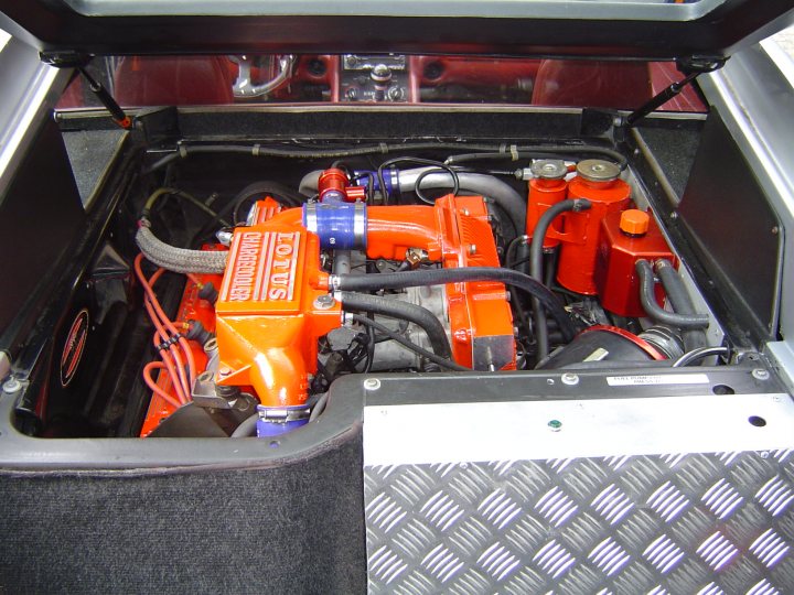 Rocker cover - Page 1 - Tuscan - PistonHeads - The image shows a close-up view of the underside of a car's engine compartment. It features a prominent red engine as the central focus. A black wire runs across the bottom of the image, possibly connecting internal components. On the left side of the image is a black rubber fender, and on the right is a silver metal plaque. The overall image gives the impression of the engine's complex inner workings.