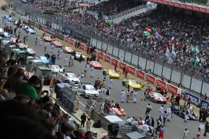 Le Mans 2011 - Page 1 - Vipers - PistonHeads - The image captures a bustling scene at a racetrack. Spectators are gathered in the stands, their attention riveted on the track below. A group of people, likely drivers, teams, or officials, are in the pits area, walking in an orderly fashion. Various individuals are standing on the sidelines, possibly preparing for the race or just passed traffic. The yellow wall of the Monaco Grand Prix circuit is visible, adding a touch of bright color to this dynamic environment. The image conveys the excitement and anticipation that commonly precede high-stakes races.