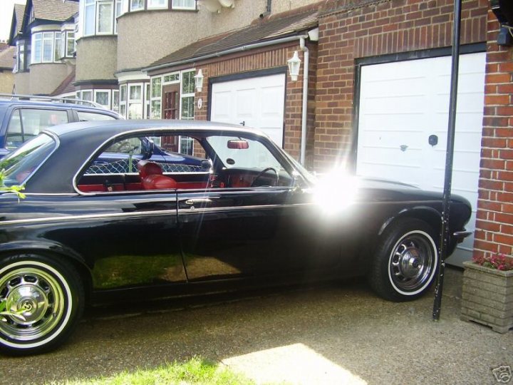 Buy Black Pistonheads - The image shows a sleek black classic car parked in front of a large, white garage door on a brick building. The car stands out with its shiny exterior and contrasting silver rims. There's a glare on the car suggesting that sunlight is present, casting light onto the hood of the vehicle. The other cars in the scene appear to be modern, while the classic car itself appears to be well-maintained and in excellent condition.