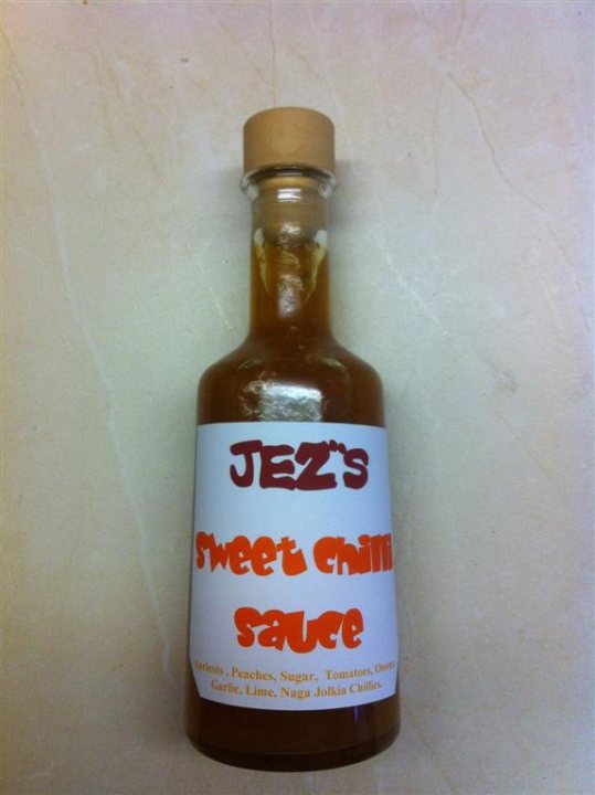 Pistonheads - The image shows a bottle labeled "Jezz's Sweet Chili sauce". The bottle appears to be made of glass and is sealed with a cork. It has a brown liquid inside, likely a sweet chili sauce, as indicated by the text on the label. The label mentions "Peach, Sugar, Tomatoes, Onions, Garlic, Lim". The background is a marble-like countertop. The image is a close-up photograph with the focus on the bottle and its label.
