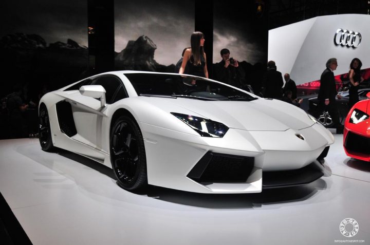 Pistonheads Geneva - The image shows a large white sports car prominently displayed at an auto show. The car is sleek and modern, featuring a low, wide stance with large, angular headlights and a distinctive front grill. The vehicle is parked on a reflective white surface, gazing towards the left of the frame, with a person visible in the background walking towards it. There are other cars in the background, and the interior wall of the show hall has a reflective, dark surface.