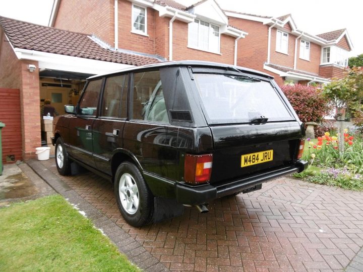 Range Rover classic - Page 14 - Land Rover - PistonHeads - The image is a photograph featuring a black Range Rover parked on a brick-paved driveway. The vehicle has a vintage or classic appearance, with a visible make or model name plate below the back glass. There are tail lights which appear to be of a type indicating the era of the vehicles' production. The setting suggests a residential area with a manicured lawn and flowers visible beside the driveway. The architecture of the houses in the background has a traditional style with red brick walls and double-hung windows. There is no visible text or distinctive branding in the image from which I can confidently confirm any make or model names of vehicles or other objects.