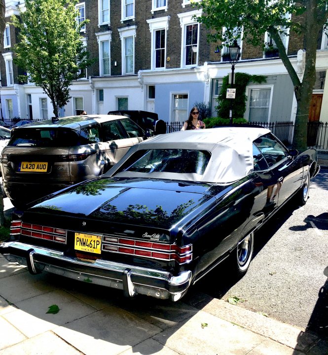 COOL CLASSIC CAR SPOTTERS POST! (Vol 3) - Page 221 - Classic Cars and Yesterday's Heroes - PistonHeads UK - The image shows a vintage black car parked on the side of a city street. It appears to be a classic muscle car, possibly a Chrysler or Dodge model given its distinctive grill and rear design. The car is parked in front of a residential building, which features multiple stories with large windows. There are also other cars visible in the background, indicating that this is a parking area. To the right of the image, there's a person partially visible, suggesting some activity happening nearby.