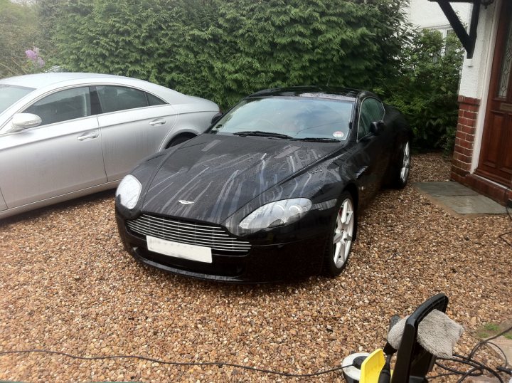 Clean Easter Pistonheads - The image shows a black sports car parked on a gravel driveway. The car is shiny, and there appears to be some water on its surface, suggesting it may have just been washed. The car is positioned next to a parked silver car, differing in size and color. To the right of the black car, there is a brick walkway leading to a house. The overall setting seems to be a residential area, and the conditions suggest it might be after a rain.