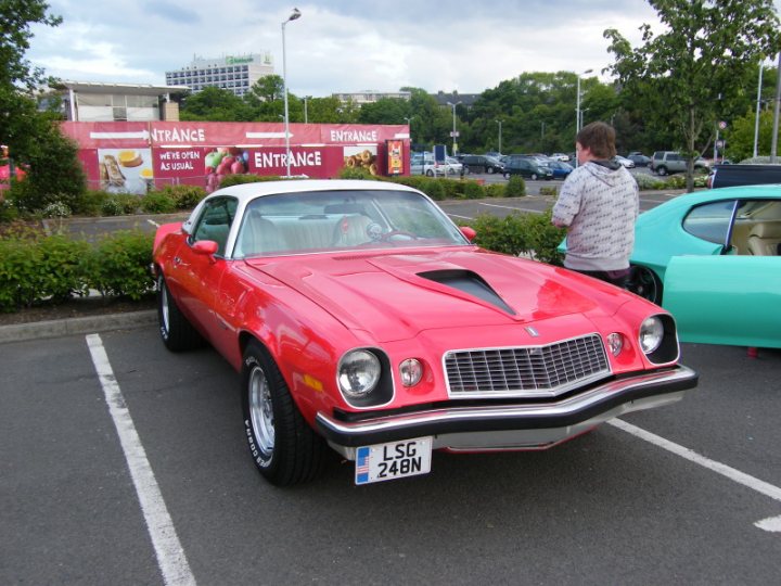 Edinburgh Cruisemeet Pistonheads - In the image, there's a parking lot where a vibrant red muscle car is parked next to a turquoise car. Behind these vehicles, you can see the side of an entrance to a shopping area shop entry sign and various other cars parked. To the right, a person is standing, observing the scene. The atmosphere appears to be calm and everyday, capturing a typical moment in a bustling shopping area.