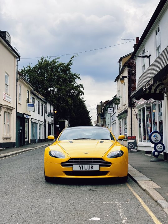 The Curfew XJ-S - V12 manual - Page 17 - Readers' Cars - PistonHeads UK - This is a color photograph showing a bright yellow Aston Martin sports car parked on the side of a narrow road. The car appears to be a two-door model with distinctive design features typical of luxury sportscars. In the background, there are buildings and a street sign, suggesting this scene takes place in an urban or suburban area. The sky is overcast, casting soft light over the scene. There are no visible texts or distinctive markings on the car that provide additional context about the location or the driver.