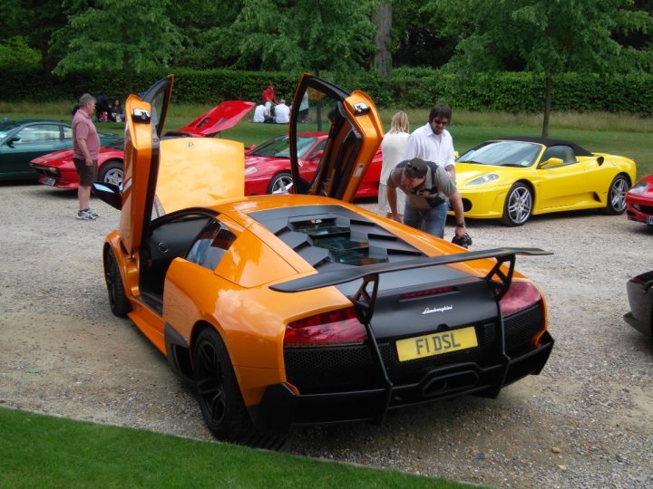 House Cliveden Pistonheads Ferrari - The image shows an expansive parking lot with a variety of cars parked on gravel. A bright orange sports car with the top panels down is the most eye-catching vehicle, its design resembling a Lamborghini, and the number "F1SDL" prominently displayed on the back. The car is positioned in such a way that the photographer peers into a slightly open driver's side window. Surrounding the car, several other individuals are visible, perhaps attendees at an auto event. The environment is lush with green trees and gardens, creating a vibrant backdrop for the cars. Notably, there are a couple of umbrellas with no people under them, possibly left behind or reserved for use later.