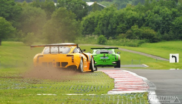 A truck driving down a road next to a forest - Pistonheads - In the photo, we see a dramatic scene from a racing event on a winding road. Two race cars, one vibrant yellow and another a sleek green, are captured in the midst of competition. The yellow car, appearing slightly faster given its position on the right side of the image and its greater distance from the camera, is leading the green car, which is a bit closer. The road, with its red and white dividers, shows a mix of dry and wet surfaces, suggesting recent or ongoing rain. Surrounding the race track, dense trees can be seen, adding a touch of the natural world to this high-speed action scene.