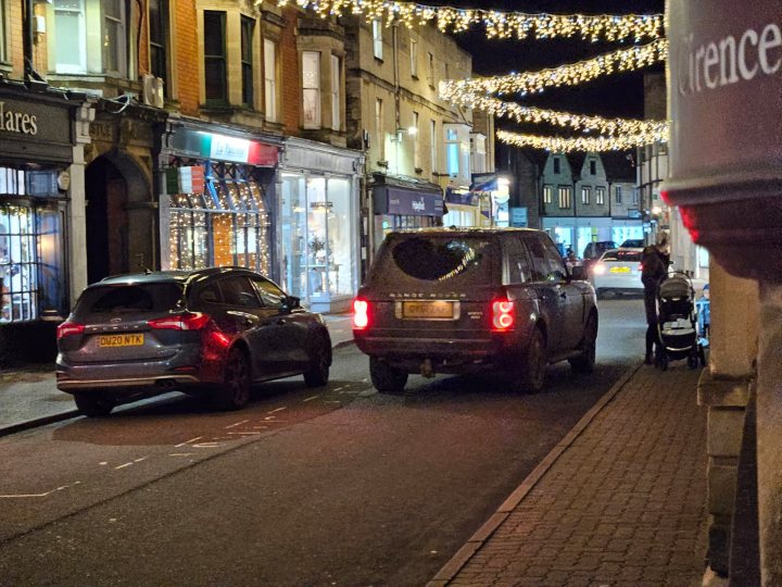 Help please identifying this plate - Page 1 - Speed, Plod & the Law - PistonHeads UK - The image depicts a quiet city street at night. Hanging lights illuminate the scene, casting a warm glow on the cobblestone road below. Parked cars line both sides of the street, their shiny surfaces reflecting the ambient light. A few pedestrians can be seen walking along the sidewalk. Storefronts with lit signs add to the festive atmosphere. The image captures the charm and tranquility of a small town or neighborhood during the holiday season.