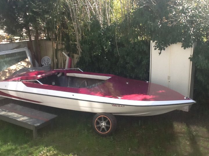 Pistonheads - The image showcases a vibrantly colored boat parked in a grassy area. The boat is predominantly maroon with a white and red stripe running down the middle. A black seat with safety rails is installed in the middle of the boat, suggesting it's for recreational use. The boat is nestled against a backdrop of lush greenery, specifically large trees and a white shed, creating a serene outdoor setting. The lighting in the picture casts a greenish hue over the entire scene, indicating it might be late afternoon or dusk. The boat's registration number is written as "53648.7".