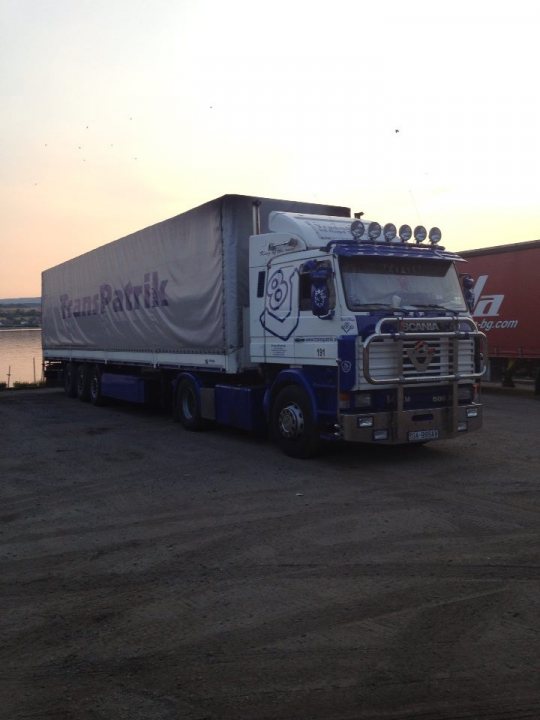 View from your cab, or outside it. - Page 22 - Commercial Break - PistonHeads - The image features a large blue and white truck parked on a gravel surface with a clear sky in the background. The truck is a semi-trailer with a white trailer attached. The cab of the truck has a custom livery with designs, including the number six. It's parked next to a large red trailer, and the backdrop gives the impression of a seaside location with a slightly curved horizon line. The style of the image is a real-life photo capturing a moment in transit.