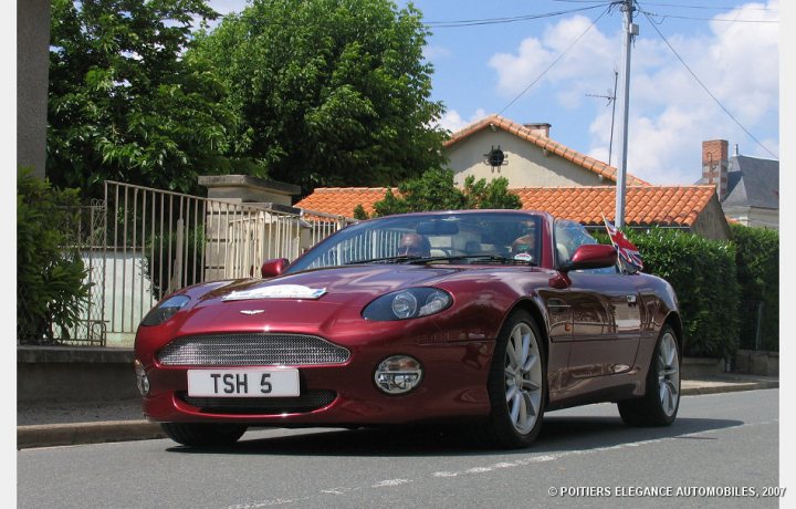 Your DB7 pictures. - Page 1 - Aston Martin - PistonHeads - The image shows a vibrant red sports car with a shiny finish, parked in an area with houses in the background. It appears to be a quiet street or a residential area, with the car in the foreground and the houses framed on the sides. The license plate of the sports car reads "TSH 5." The sky is clear, suggesting it might be a pleasant day. The car seems to be in pristine condition, possibly indicating it is well taken care of.