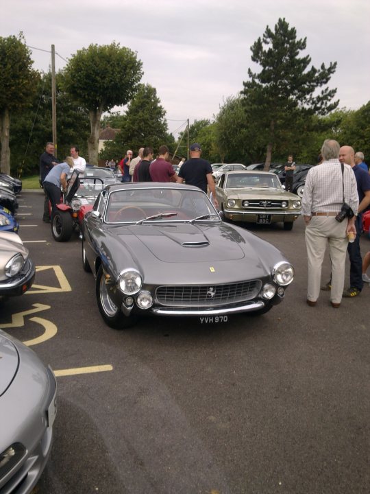 Pistonheads - The image depicts a gathering of enthusiasts in a parking lot, focusing on a row of classic cars. Two distinct models are featured prominently: a Ferrari convertible with a black interior and a white Monaco, both boasting their iconic badges. Multiple individuals, some carrying handbags and backpacks, are milling around and engaging with the vehicles, appreciating their design and history. The setting is serene with a lush backdrop of green trees, creating a contrast with the gleaming cars and the concrete ground.