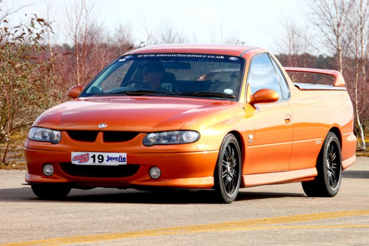 The image shows an orange, four-door coupe parked on what appears to be a street, with the vehicle facing towards the left side of the frame. On the license plate, the text "19 Jevs" can be seen. In the background, there are trees, indicating a suburban or residential area. The car seems to be in good condition, as it is photographed in the daylight. The photo is taken from the front, providing a clear view of the car's exterior, including the hood and the front wheels.