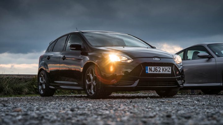 A car parked next to a parking meter - Pistonheads - The image shows a black four-door Ford Fiesta car parked on a gravel surface on what appears to be either a cloudy or overcast day. The car is facing the camera, highlighting its front and headlights. The sun is setting in the background, casting a gentle golden glow on the horizon. Another car can be seen parked slightly beside the Fiesta in the distance, capturing the elements of a street or parking area during dusk.