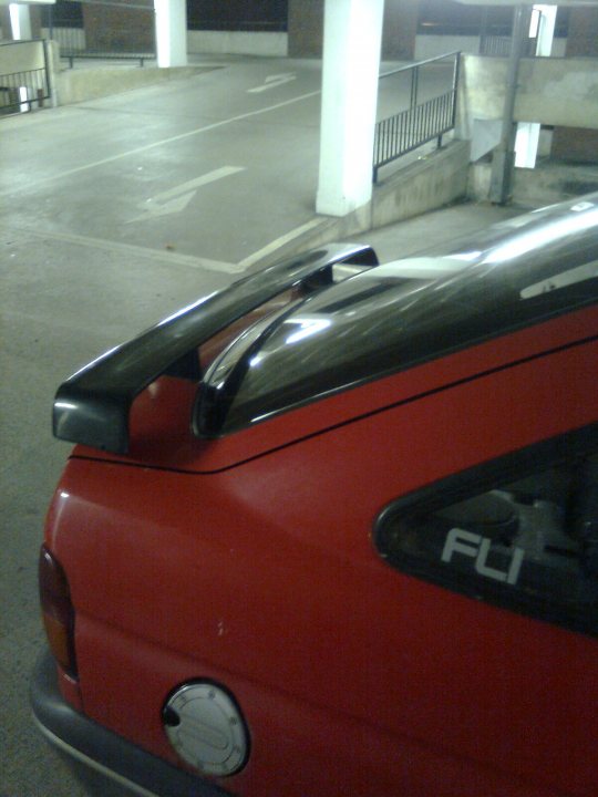 Escort Pistonheads - The image features a close-up of a red sports car parked in a mostly vacant parking lot in a public area during the night. The car's front left side is visible, with the left rear fender, the black spoiler on the roof, and the car's emblem being most prominent. The body of the car has a glossy finish, reflecting some ambient light. The parking lot has some visible streetlights, adding to the coverage of the area, and providing areas of harsh and softer light which create depth in the photograph.