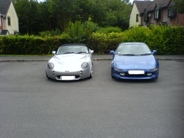 She's a write off. - Page 2 - Jap Chat - PistonHeads - The image depicts two sports cars parked side-by-side in an outdoor parking lot. The car on the left is a silver Porsche Boxster, while the car on the right is a blue Mazda Miata. Both cars have a sleek, aerodynamic design, indicative of their sporty nature. In the background, there is a row of brick houses with lush trees and bushes, implying a residential area.